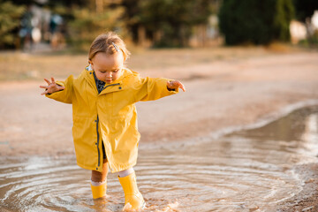 Cute baby girl 3-4 wearing yellow raincoat and rubber boots walking in puddle outdoors. Autumn...