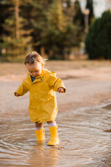 Cute baby girl 3-4 wearing yellow raincoat and rubber boots walking in puddle outdoors. Autumn season. Childhood.