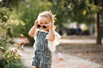 Smiling cute baby girl 2-3 year old wearing sun glasses and stylish summer clothes in park outdoors. Looking at camera. Childhood.