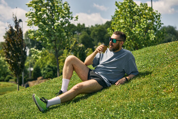 Man in midlife unwinding post-run on grassy hill with water bottle beneath sunny blue sky. Concept...