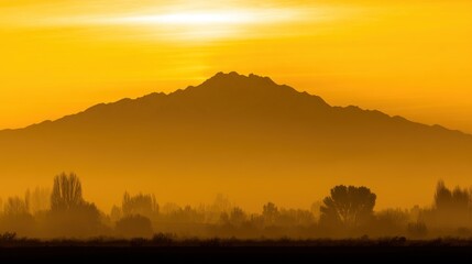 Obraz premium Serene mountain landscape at sunrise or sunset with warm golden-orange sky, silhouetted peaks, misty foreground, and hazy silhouettes of trees and shrubs.