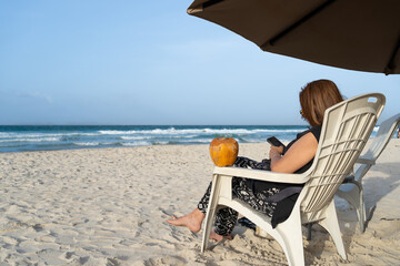 La señora de la tercera edad está disfrutando la playa sentada en su silla, con su sombrilla, su...