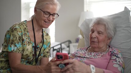 A compassionate nurse engages with an elderly woman by sharing photos and memories on a smartphone during a visit at a senior living facility.