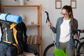 woman hiker packing backpack for mountain hike holding nordic walking poles at home.