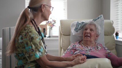 A nurse holds hand of an elderly woman while engaging her in friendly conversation in a senior living facility. Both appear happy and engaged.
