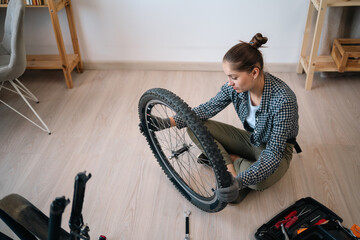 Young woman repairing servicing her bicycle holding bike wheel sitting on floor at home