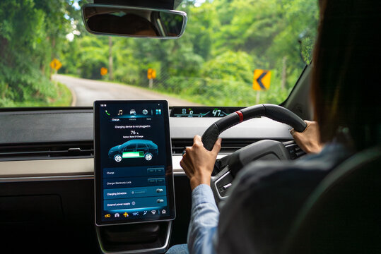 Asian woman driver electric car along a scenic winding road, surrounded by lush greenery. The digital touchscreen displays the electric car battery level and charging options. Future energy concept.