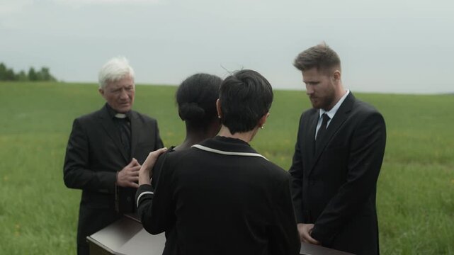 Mature Caucasian priest in black soutane saying funeral speech in front of diverse sorrowing people standing by wooden casket at funeral ceremony outdoors