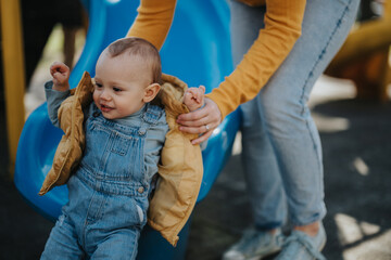 A mother supports her cheerful toddler as they enjoy time together at an outdoor playground,...