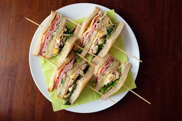 Piatto con tramezzini farciti assortiti: prosciutto, formaggio, pomodoro e pollo. Panini morbidi serviti per buffet, brunch o aperitivo. Vista dall’alto su sfondo in legno.