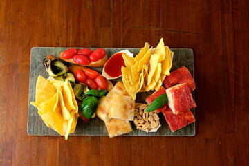Tagliere misto per aperitivo con focaccia, bruschette, pomodorini, verdure grigliate, patatine, arachidi, salsa rossa e pizza al pomodoro. Vista dall’alto su tavolo in legno.