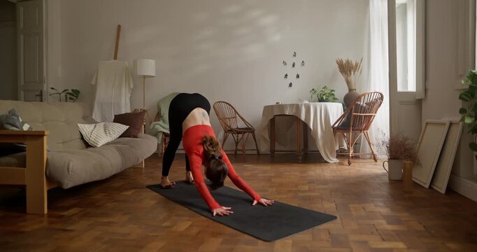 Girl in orange top doing yoga In a pose Eka Pada Adho Mukha Svanasana on Sports black rug at home. Girl in orange sweater lifts one leg up while doing yoga at home