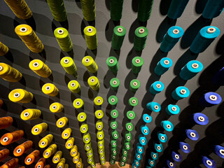 Organized wall display of colorful thread spools arranged in rainbow gradient from red to blue. Textile manufacturing or sewing supply storage showing vibrant geometric pattern background.