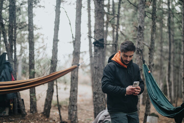 A young man is engrossed in using his smart phone while standing in a forest campsite. Hammocks are...