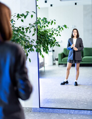 Fototapeta premium Stylish woman poses in front of a mirror. A fashionable woman stands in a modern interior, adjusting her blazer while reflecting on her look in a large mirror.