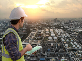 Male engineer wearing safety gear uses a digital tablet while overseeing a large oil refinery...