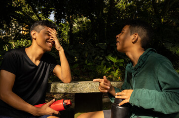 Two young Latinos sitting and talking happily surrounded by nature, enjoying the morning sun