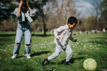 A woman enjoys a playful moment with a boy who is chasing a ball in a sunny park.