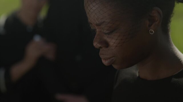 Tilt up slowmo of young Black widow wearing mourning veil gently touching wooden casket and grieving loss at outdoor funeral ceremony among family and friends