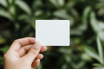 Person holding a blank white card in front of green foliage. Minimalist design, perfect for messages, promotions or notes in a natural setting.