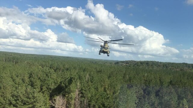 A CH-47 Chinook tandem-rotor helicopter hovers above a dense forest beneath dramatic cumulus clouds. Renowned for its heavy-lift capabilities, the Chinook is used for cargo transport, troop movement, 