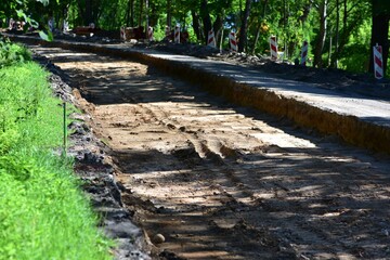 Dug-out road under reconstruction in green forested area. Graded dirt surface and tire marks show...
