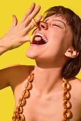 Joyful woman biting bagel rings from fingers in surreal yellow snack fashion. Concept of edible...