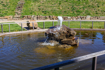 Public park pond with a fountain, water gushing from water spout over huge stone, surrounded by railing, village street in background, sunny day in Gulpen, South Limburg, Netherlands
