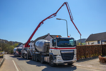 Parked cement trucks and crane with concrete discharge hose on provincial street, supply cement to construction site in background on a sunny day in Gulpen, South Limburg, the Netherlands