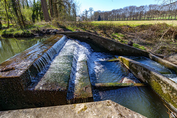 Concrete waterfall steps of small dam with water flowing into Gulp River, meadow with trees and green grass in background, sunny day in Gulpen, South Limburg, Netherlands