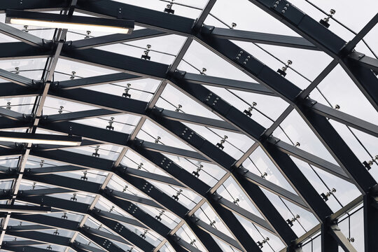 Modern steel and glass roof. Geometric structure with arched beams. Transparent ceiling design in public space. Industrial metal framework. Architectural engineering with clear panels.
