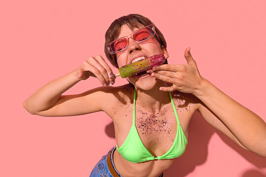 Excited woman in green bikini biting glitter-covered popsicle with playful expression. Concept of surreal food fun, pop summer styling, bold youth visuals, sweet chaos celebration.