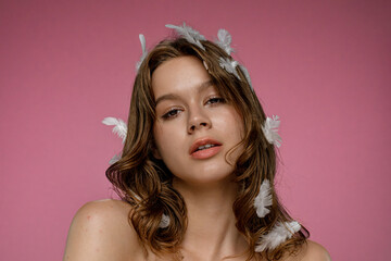 Female model with natural beauty posing with white feathers in her hair on pink background