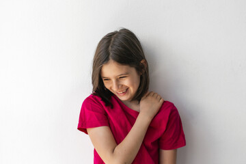 Obraz premium Shy smiling girl looking down with hand on shoulder against white wall, concept of childhood innocence and warmth