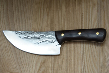 Close-up of a handcrafted chef knife with a textured steel blade and dark wooden handle placed on a wooden surface. Professional culinary tool for cooking and food preparation.