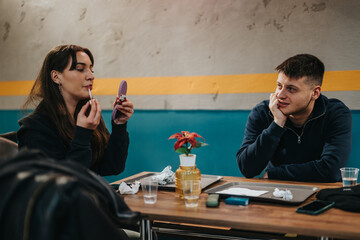 Two friends spending time at a modern cafe, with one applying makeup while the other looks on,...