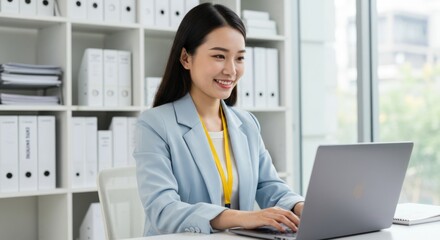 A smiling young woman in a light blue blazer sits at a desk in a modern office, using a laptop.  She has long dark hair and appears confident and professional. The office is bright and well-organized