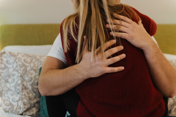Close-up of intimate embrace between two people sitting on bed, showing emotional support through...