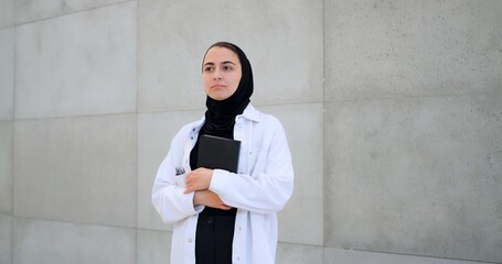 Medical professional wearing hijab and white coat standing near gray wall, holding medical textbook...