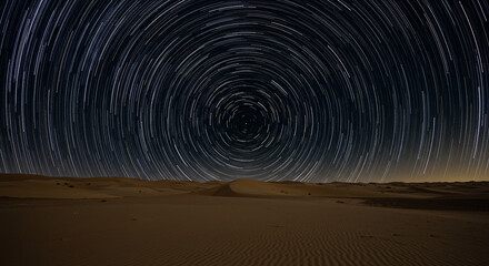 Star trails over a desert landscape with visible sand dunes and a dark night sky above the horizon