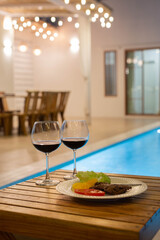 A cozy, romantic poolside dinner setting with two glasses of red wine and a plated steak dish.