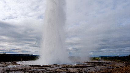 Strokkur geyser erupts powerfully into the air, surrounded by a rugged landscape in Iceland. Visitors marvel at this natural wonder, showcasing the beauty of geothermal activity in the region.
