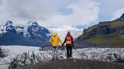 A couple wearing vibrant jackets stands on the rugged terrain, hand in hand, gazing at the...