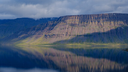 Stunning cliffs create dramatic reflections in the calm waters of a fjord in Iceland. Soft light illuminates the rocky terrain, enhancing the serene atmosphere of this breathtaking landscape.