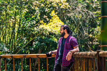 Man in plaid shirt with tied hair holding camera and gazing at tropical forest from wooden platform...