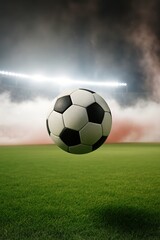 Black and white soccer ball suspended mid-air on a green field under dramatic stadium lighting, with fog and a hint of red flare in the background, capturing an intense game atmosphere.
