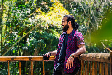 Man in plaid shirt with tied hair holding camera and gazing at tropical forest from wooden railing...