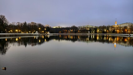 A river runs through the city in concrete banks with buildings and trees standing on them. In the evening twilight, the illumination of buildings and lanterns on the embankment are lit. Cloudy