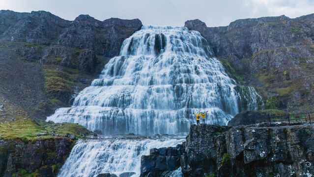 A breathtaking waterfall tumbles down rugged cliffs, surrounded by stunning landscapes. A couple stands triumphantly near the base, embracing the wild beauty of Dynjandi waterfall, Westfjord, Iceland