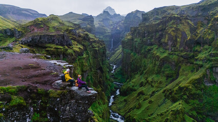 Adventurers sit on the edge of a rocky outcrop in Mulagljufur Canyon, surrounded by lush greenery...
